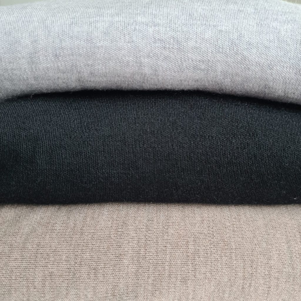 Merinovillan hoito. Merinovilla kangaspakka, jossa on päällekkäin kolme eriväristä kangasta. Ylimpänä vaalea harmaa, keskellä musta ja alimpana taupe.